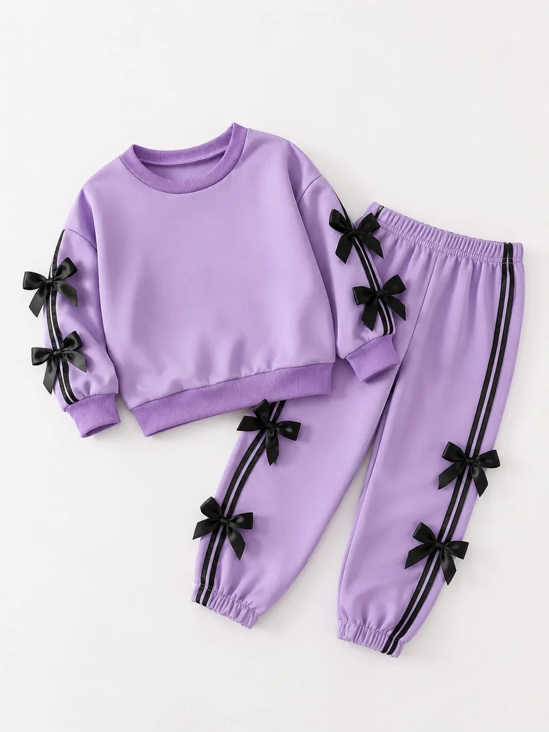 A children's two-piece tracksuit in light purple, featuring a long-sleeve sweatshirt with black ribbon bow details on the sleeves and matching jogger pants with black side stripes and bow accents, laid flat on a white background.