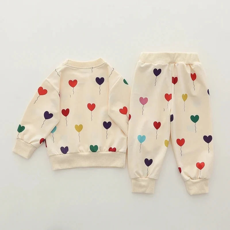 A two-piece baby clothing set featuring a cream-colored sweatshirt and matching jogger pants, both decorated with a colorful pattern of small heart-shaped balloons in red, purple, green, yellow, and blue.