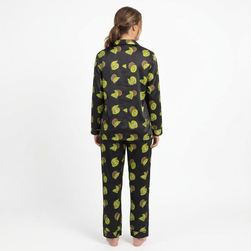 A woman viewed from behind wearing a matching long-sleeve pajama set with a black satin-like base and a repeating avocado print in green and brown, set against a plain white studio background.