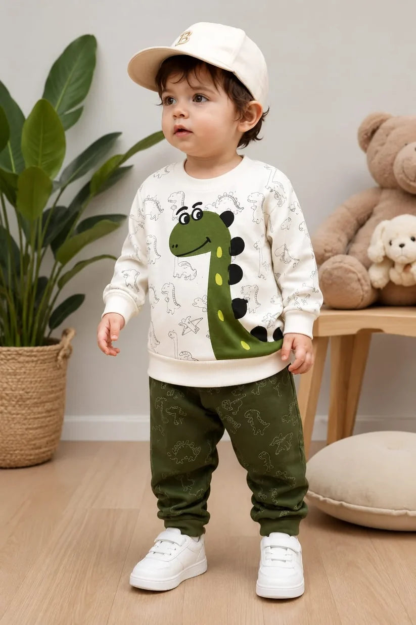 A toddler stands indoors wearing a cream long-sleeve sweatshirt with a green dinosaur graphic, olive green patterned pants, white sneakers, and a cream baseball cap. The room has a plant, a wooden table with teddy bears, and a light-colored wall.