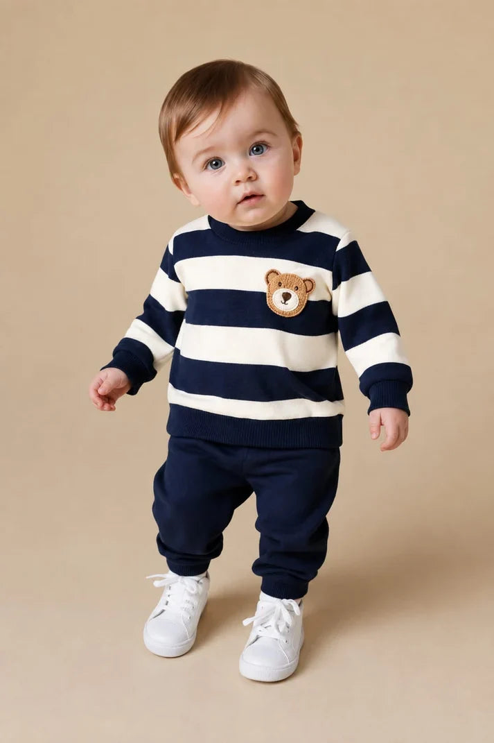 A baby standing against a beige studio background, wearing a navy and cream striped long-sleeve top with a teddy bear face on the chest, navy pants, and white sneakers.