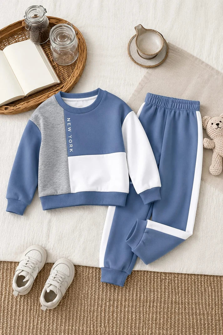 A children's two-piece sweatshirt and jogger set laid flat on a neutral fabric background with styled props. The sweatshirt is blue, gray, and white with 'NEW YORK' printed vertically on the chest.