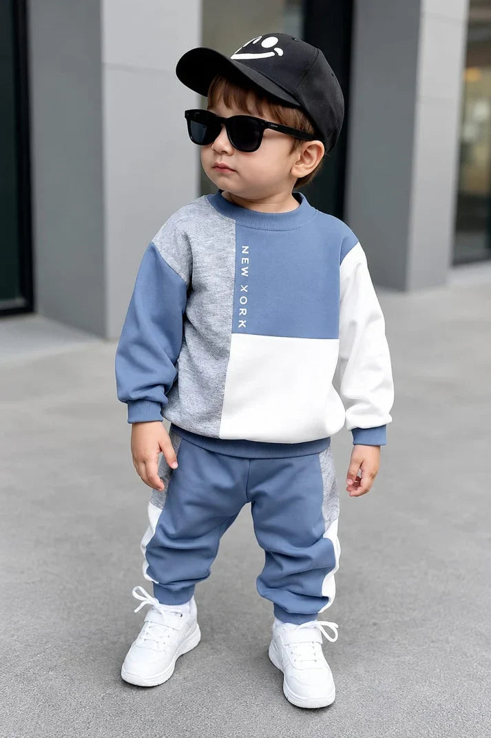 A toddler wearing a matching two-piece tracksuit set, featuring a color-blocked sweatshirt in blue, gray, and white with 'NEW YORK' printed vertically, matching blue jogger pants with white side stripes, a black baseball cap with a white smiley face, and black sunglasses, standing on a gray concrete surface.
