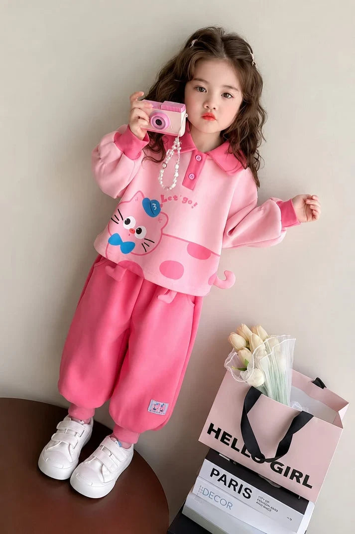 A young girl modeling a pink two-piece outfit consisting of a long-sleeve top with a cartoon cat face graphic and matching pink pants, standing next to a shopping bag and books.