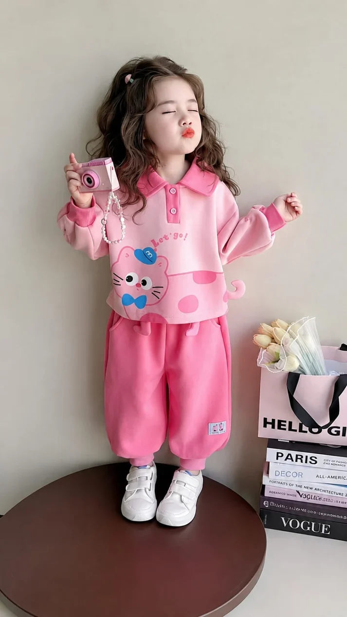 A young girl modeling a pink two-piece pajama set consisting of a long-sleeve polo shirt with a cartoon cat graphic and matching sweatpants, standing on a round wooden platform.