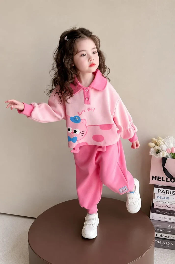 A young girl wearing a pink two-piece outfit with a cartoon cat graphic on the sweatshirt and matching pink sweatpants, posing on a round brown pedestal in a studio setting.
