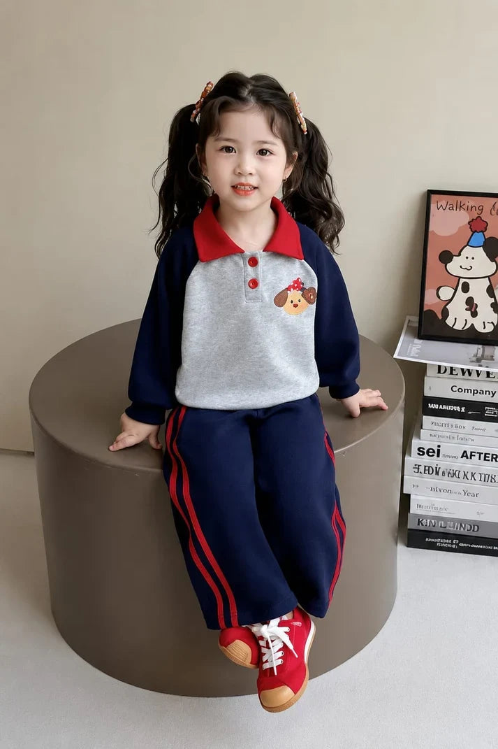 A young girl with dark curly hair in pigtails sits on a round brown stool, wearing a two-piece athletic tracksuit. The set features a light gray and navy blue long-sleeve top with a red collar and a dog graphic on the chest, paired with matching navy blue pants that have red side stripes.