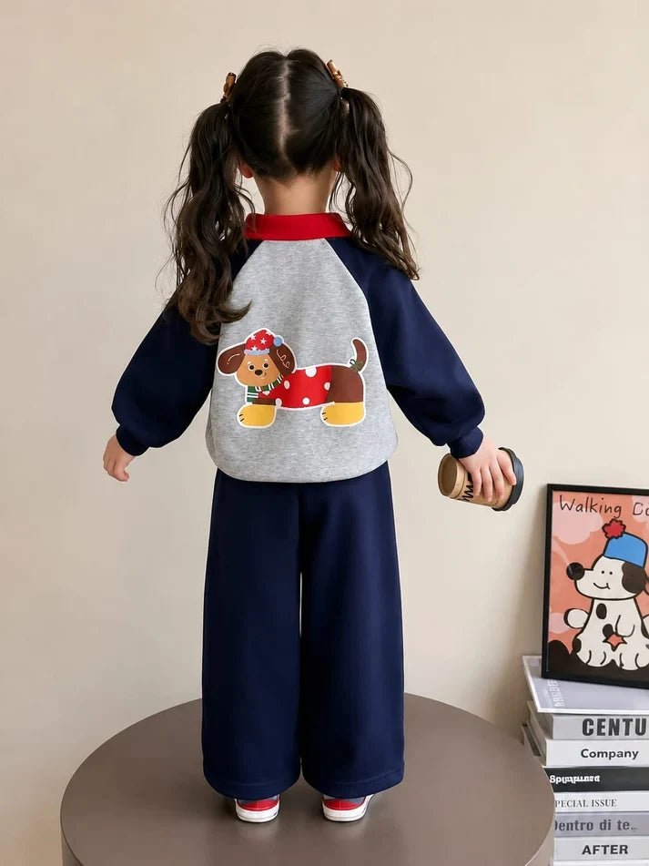 A young girl is shown from behind wearing a two-piece children's outfit. The set consists of a gray and navy blue raglan-style sweatshirt with a cartoon dachshund graphic on the back, and matching navy blue wide-leg pants.