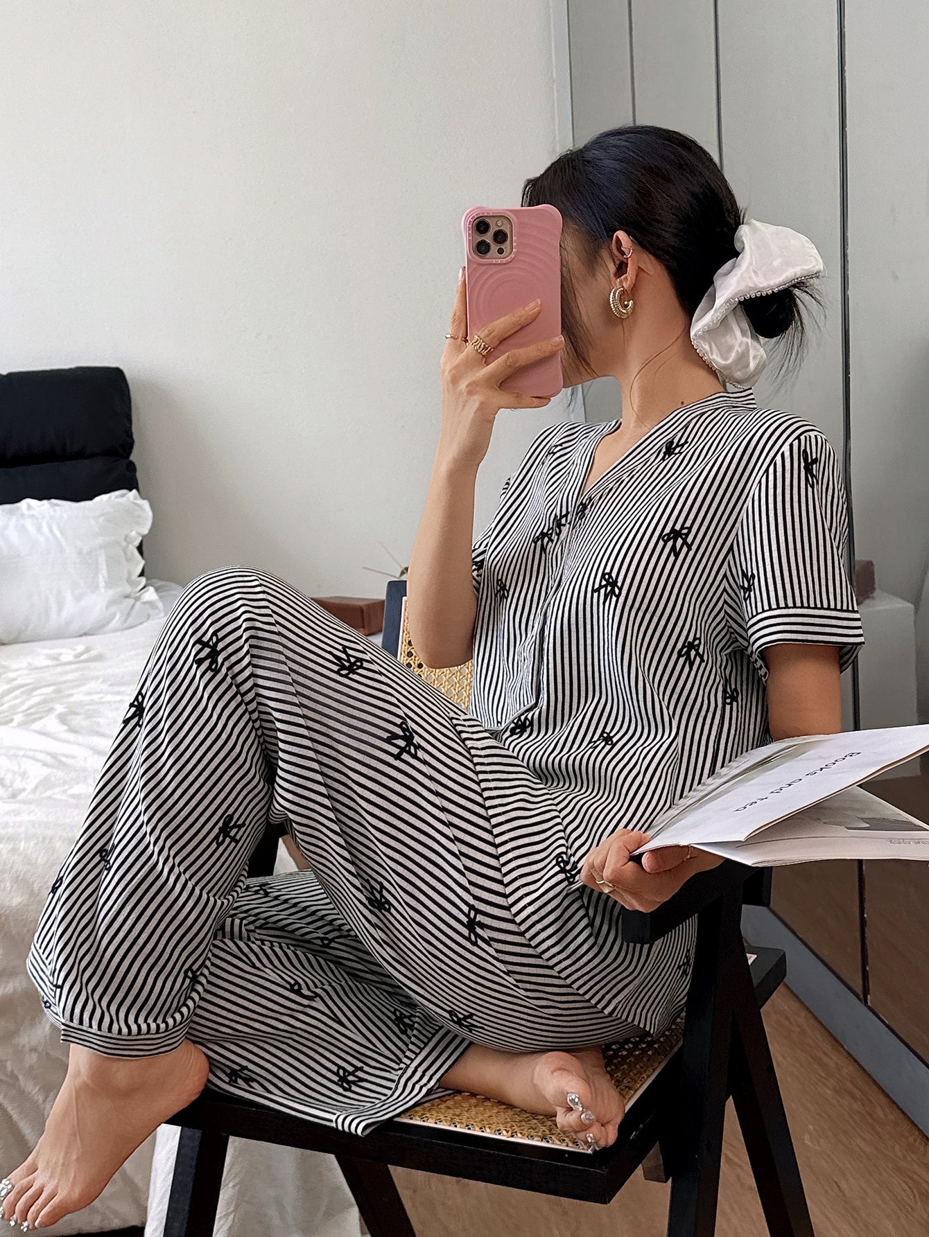 A woman wearing a black and white striped pajama set with small embroidered motifs, sitting on a stool in a bedroom while taking a mirror selfie with her phone.