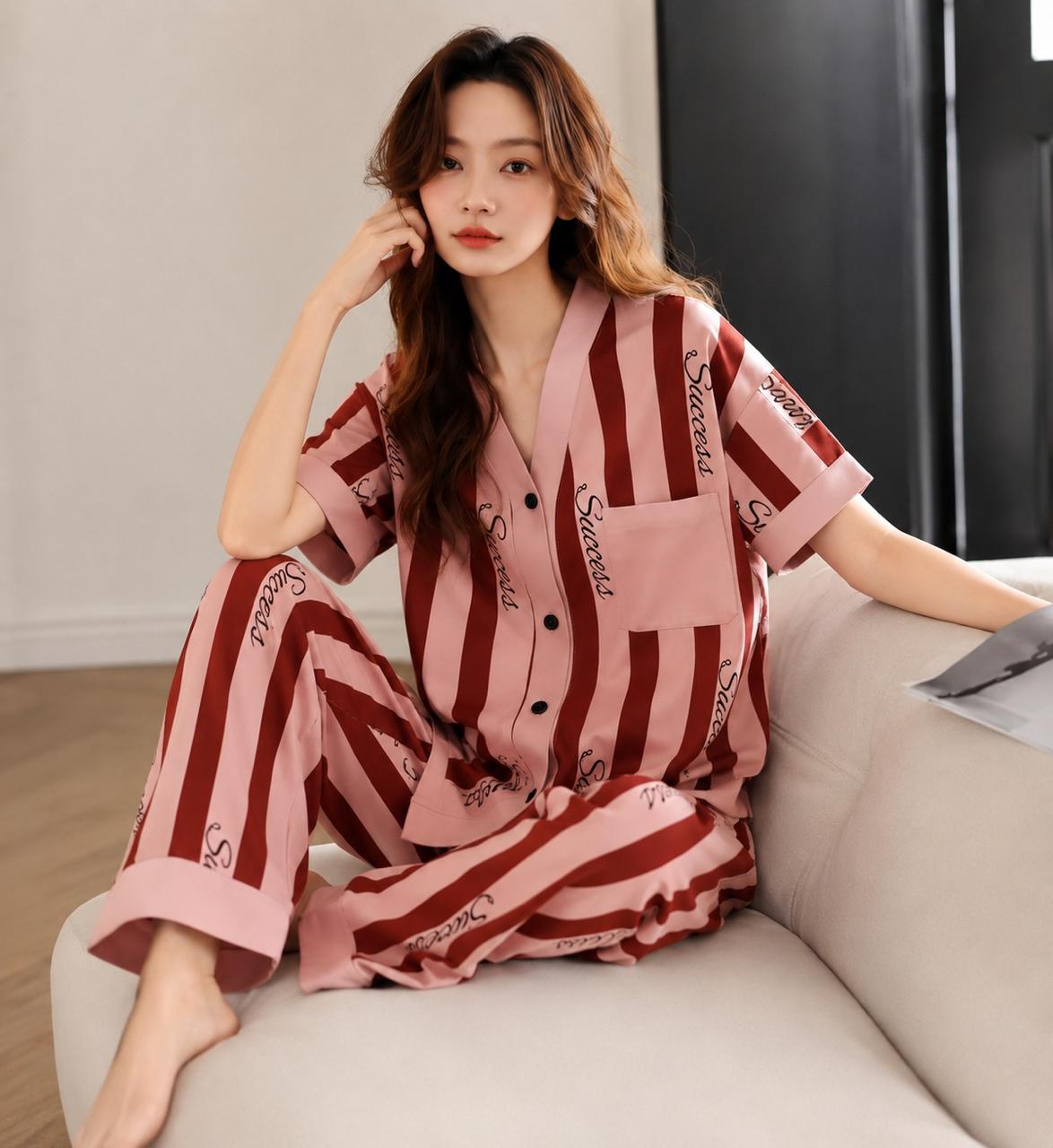 A woman sits on a light beige sofa wearing a pink and dark red striped pajama set with black script text details. The room has a neutral background with a light wall and dark paneling.