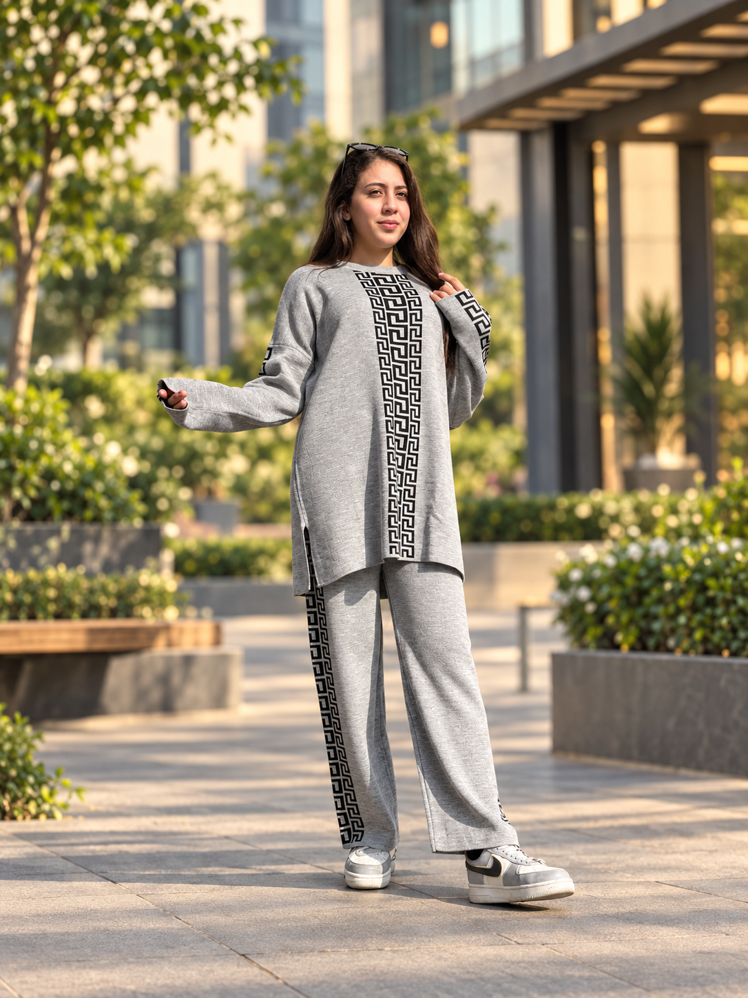 A woman stands outdoors on a paved walkway wearing a light gray two-piece lounge set with black geometric patterned trim down the front and along the sleeves and pants. The background shows modern glass buildings and greenery.