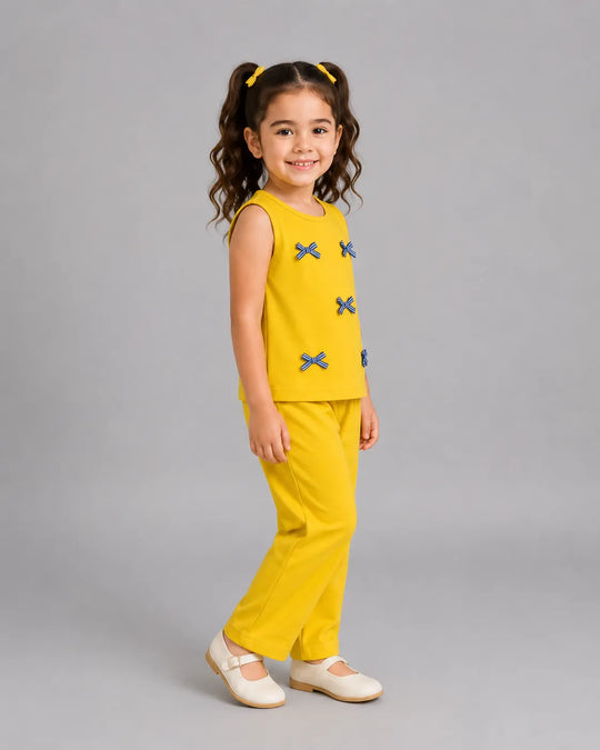 A young girl with curly brown hair in pigtails stands in profile wearing a bright yellow sleeveless top and matching yellow pants, both decorated with small blue bow motifs. She is wearing white ballet flats against a solid gray studio background.
