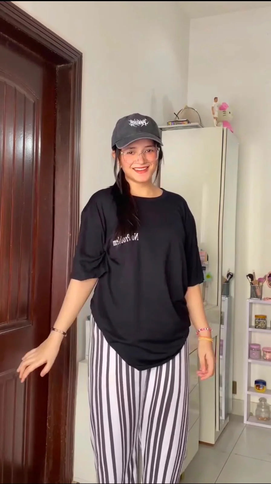 A woman wearing a black oversized t-shirt with white text, black-and-white vertically striped pants, and a black baseball cap, standing in a room next to a dark brown wooden door.